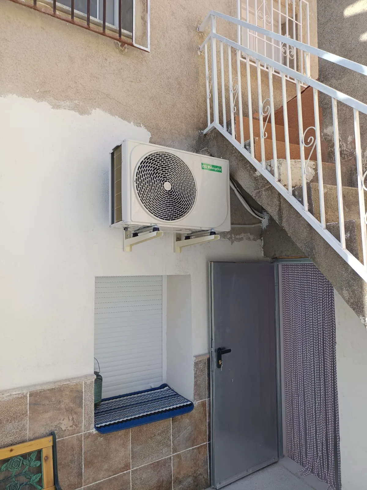 Unidad exterior de aire acondicionado instalada en la pared de un patio, junto a la escalera.